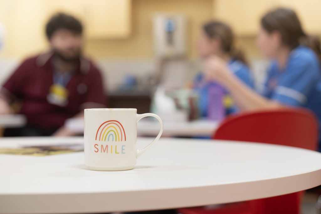 In the foreground, a mug with the word 'Smile' on it, and a child's drawing of a rainbow. In the background, some out-of-focus staff members chatting over coffee.